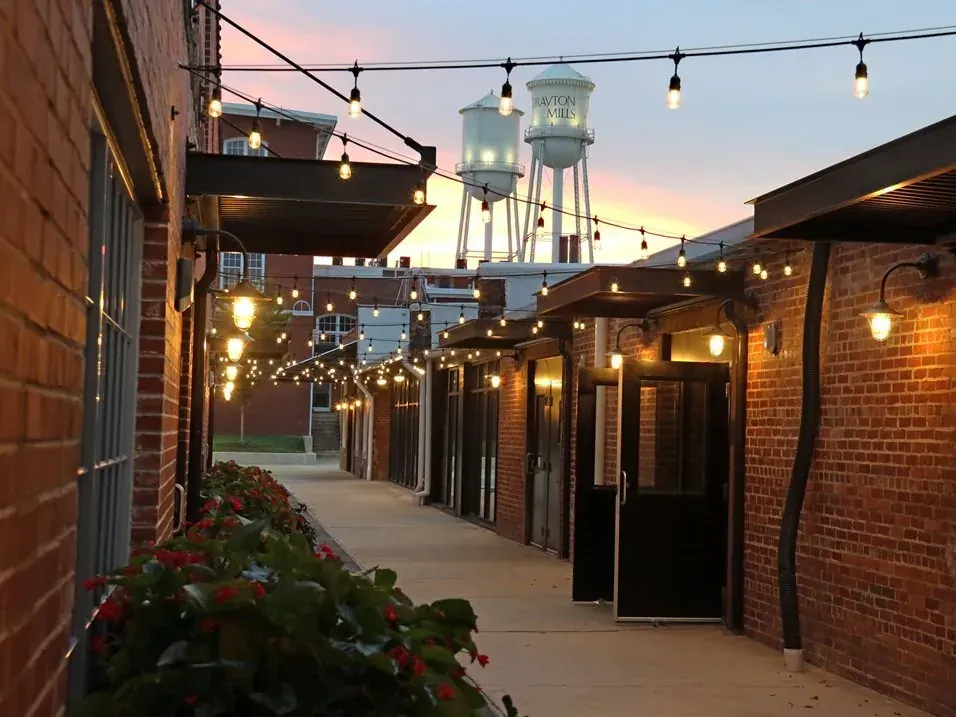 String lights over the brick walkway of Drayton Mills Marketplace at dusk, with the historic water towers behind